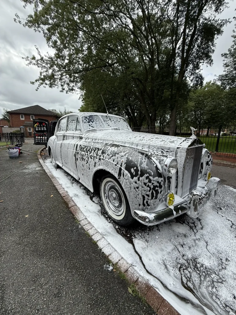 Classic Rolls-Royce covered in wash foam beside a tree-lined road